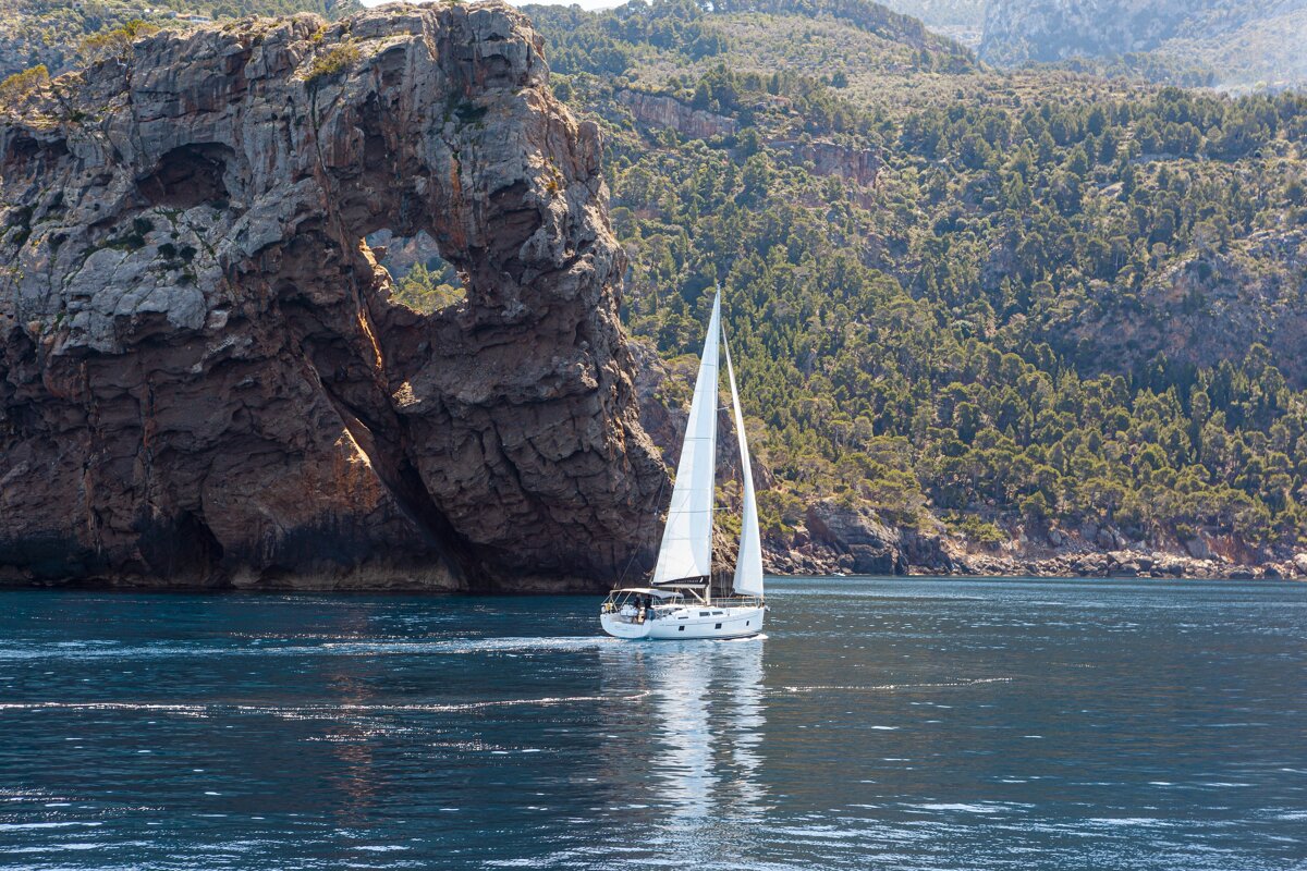 A white sailboat glides across blue water next to a massive rocky cliff with a natural arch.