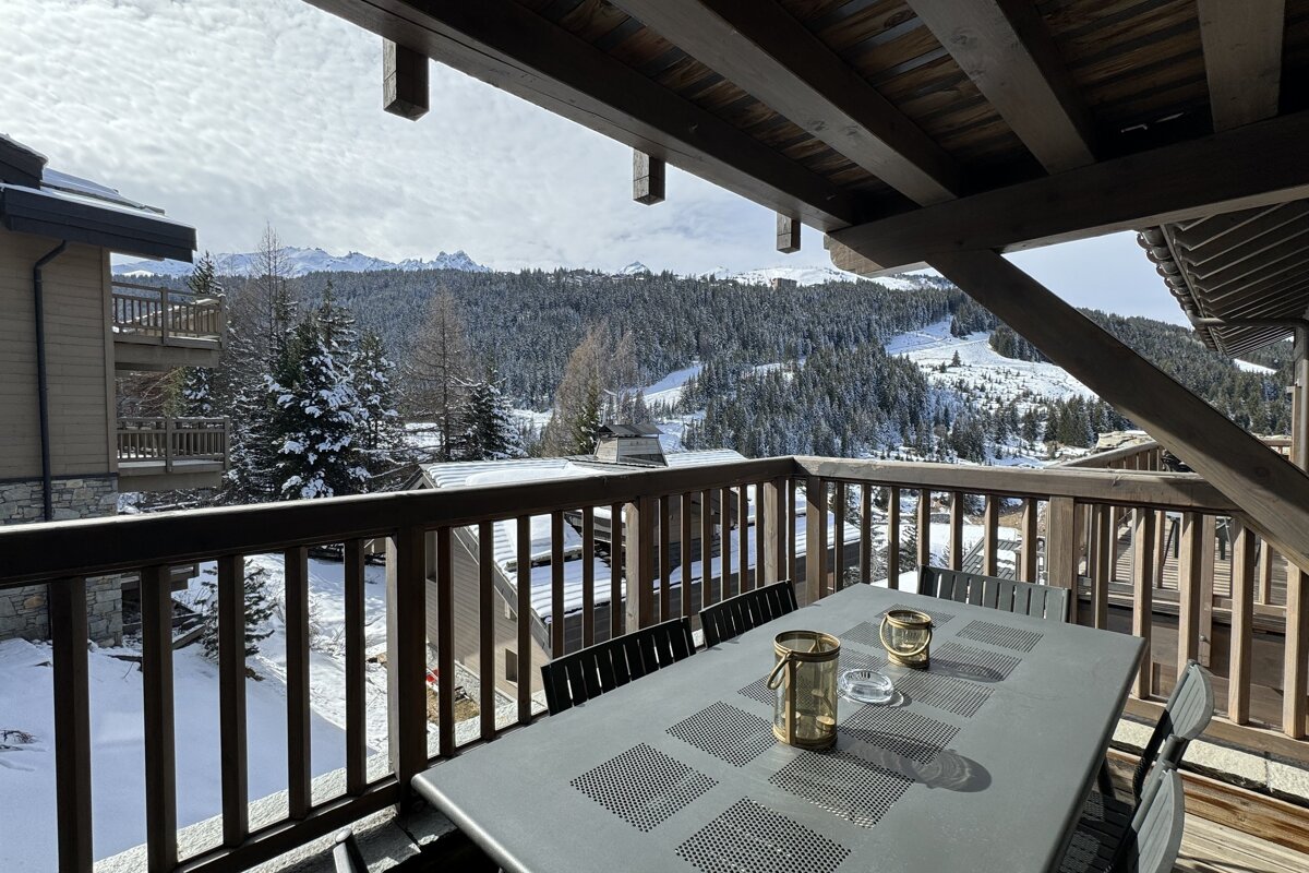 A cozy balcony with outdoor seating overlooks a vast, snow-covered mountain landscape, dense pine forests, and distant ski slopes under a winter sky.