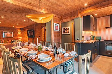 A stylish, modern dining and kitchen area with warm wooden walls and ceiling. The dining table is set, next to a black kitchen with a pink striped accent wall.