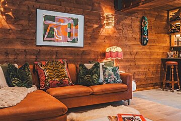 A cozy room features warm wood-paneled walls, an orange L-shaped sofa with vibrant cushions, abstract art, a unique lamp, and a home bar.