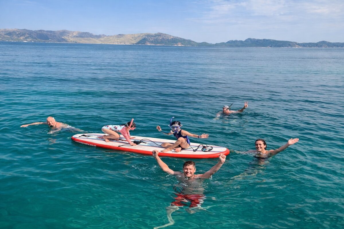 A joyful group of people, including kids on a paddleboard, swim and snorkel in clear blue ocean waters with mountains in the distance.