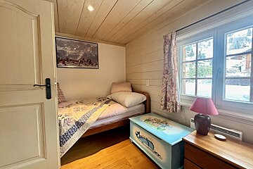 A small, rustic bedroom with a single bed, wooden interior, a window with a snowy view, and a colorful painted chest.