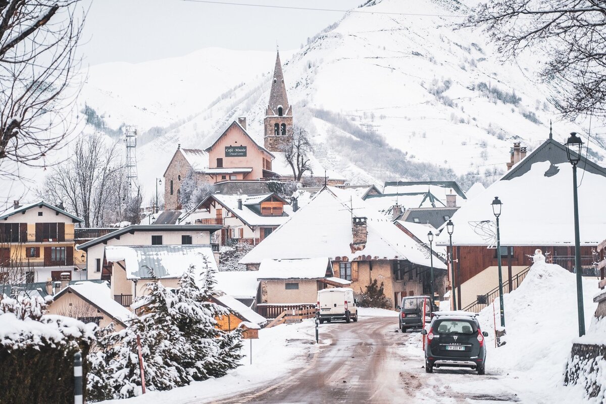 A charming, snow-covered mountain village with a church steeple and houses nestled against a white mountain, a road winding through it.