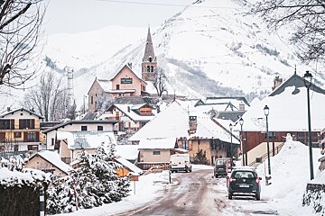 A charming, snow-covered mountain village with a church steeple and houses nestled against a white mountain, a road winding through it.