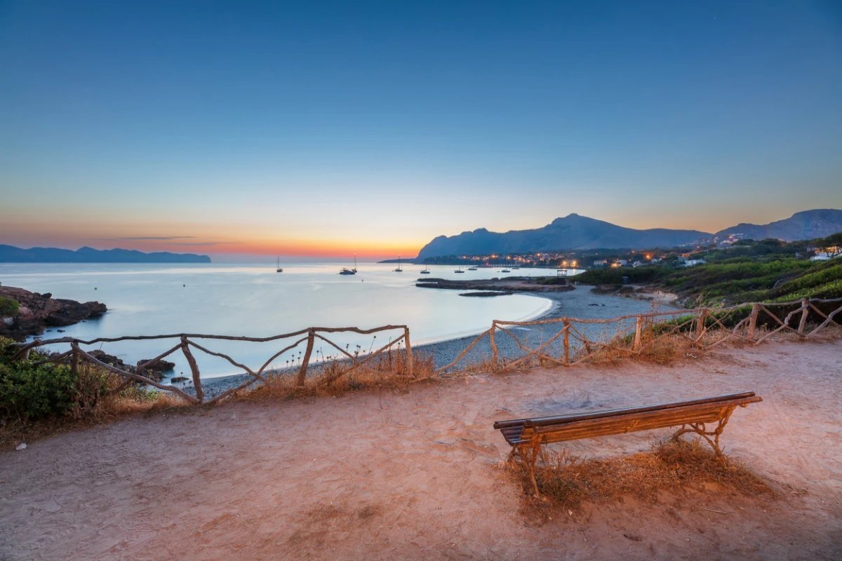 Dusk view over a calm bay with boats, distant town lights, and mountains, seen from a viewpoint with a rustic fence and a wooden bench.