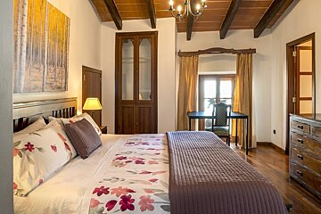 A cozy bedroom with a large bed, wooden furniture, a desk by a bright window with curtains, beamed ceiling, and nature-themed art.