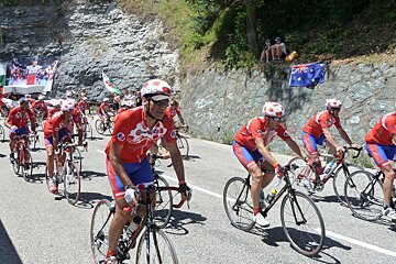 A group of cyclists are riding down a road with flags behind them