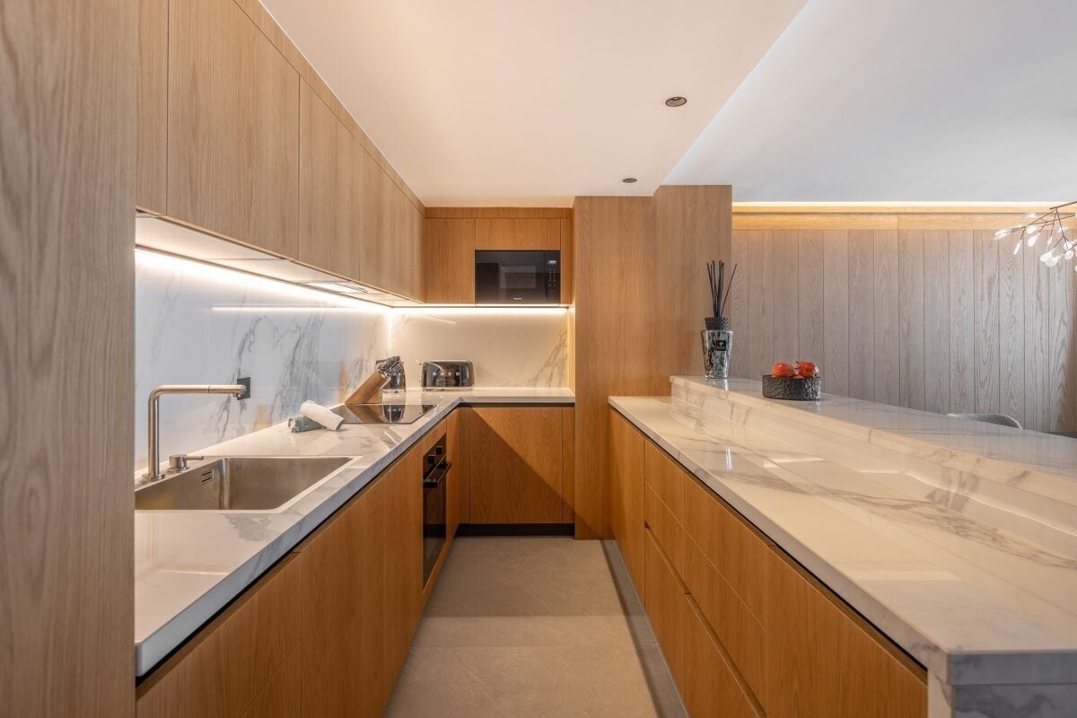 A modern kitchen with light wood cabinets, white marble countertops, and undercabinet lighting. Features a sink, stovetop, microwave, and a bar area.
