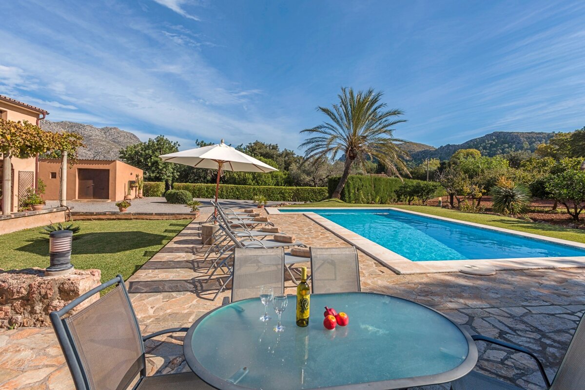 A sunny villa patio with wine, fruit, and a refreshing pool. Lounge chairs, palm trees, and mountains complete the idyllic scene under a blue sky.