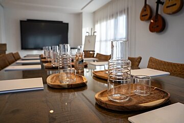A modern meeting room with a long table set with water carafes, glasses, and notepads. A large TV screen and mandolins decorate the walls.