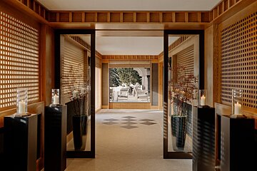 A serene, wood-paneled hallway with intricate lattice screens and sliding mirror doors, opening to a snowy outdoor patio. Candles glow on pedestals.
