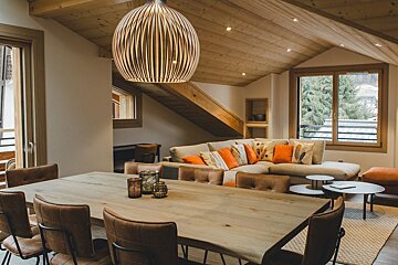 A cozy, modern open-plan living/dining room features a large wooden table, sofa with orange cushions, wooden ceiling, and a view of trees.