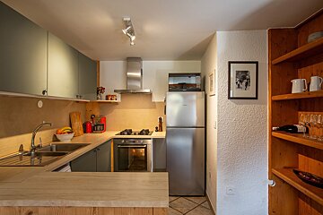 A modern kitchen features gray-green cabinets, wooden countertops, stainless steel appliances including a fridge and oven, a sink, and open wooden shelves.