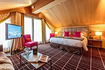 Luxurious attic hotel room with a large bed, pink accents, wooden ceiling, patterned carpet, and a TV showing snowy mountains.