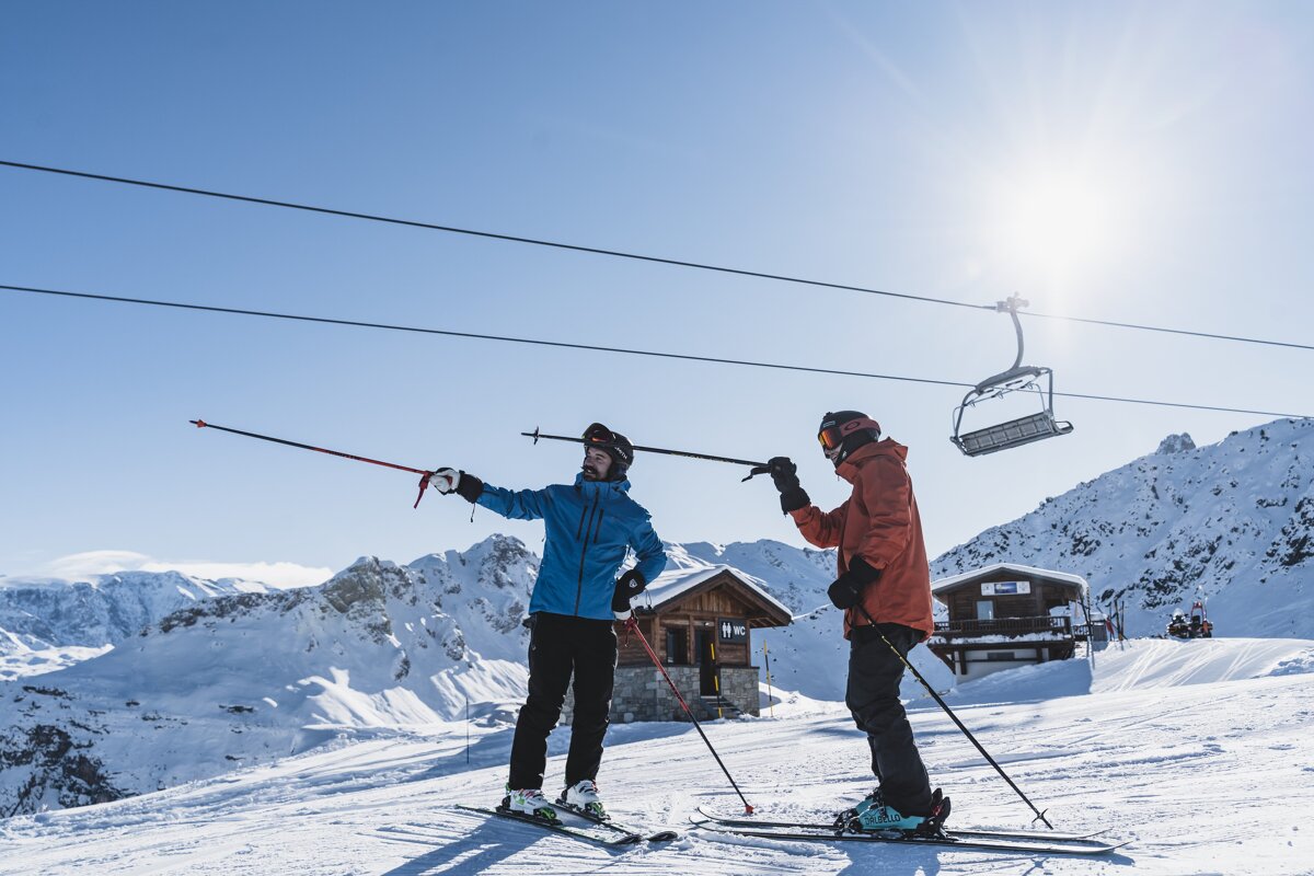 Two skiers on a sunny snowy mountain slope, one pointing with a pole. A ski lift and small wooden cabins are visible, surrounded by snow-covered peaks.