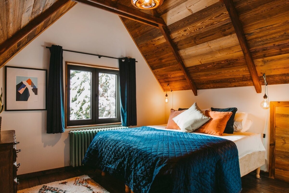A cozy attic bedroom features a rustic wooden ceiling, a blue quilted bed, and a window offering a snowy view. Warm lighting enhances the inviting ambiance.