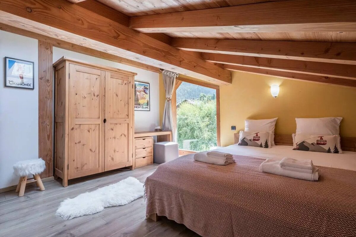 An inviting bedroom with exposed wooden beams, a large bed with mountain-themed pillows, a pine wardrobe, and a scenic mountain view from a big window.