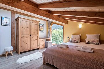 An inviting bedroom with exposed wooden beams, a large bed with mountain-themed pillows, a pine wardrobe, and a scenic mountain view from a big window.