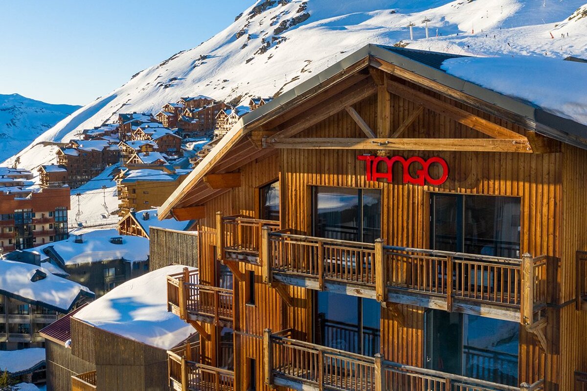A sunny ski resort view featuring a large wooden building with balconies and a red 
