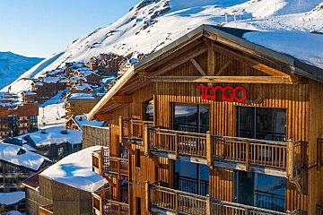 A sunny ski resort view featuring a large wooden building with balconies and a red 