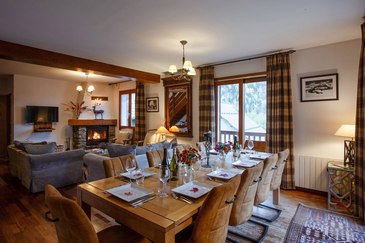 A cozy chalet living-dining room with a set wooden table, warm fireplace, comfy sofas, and large windows revealing a scenic outdoor view.