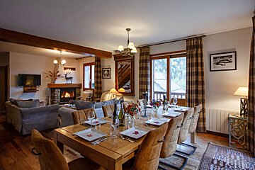A cozy chalet living-dining room with a set wooden table, warm fireplace, comfy sofas, and large windows revealing a scenic outdoor view.