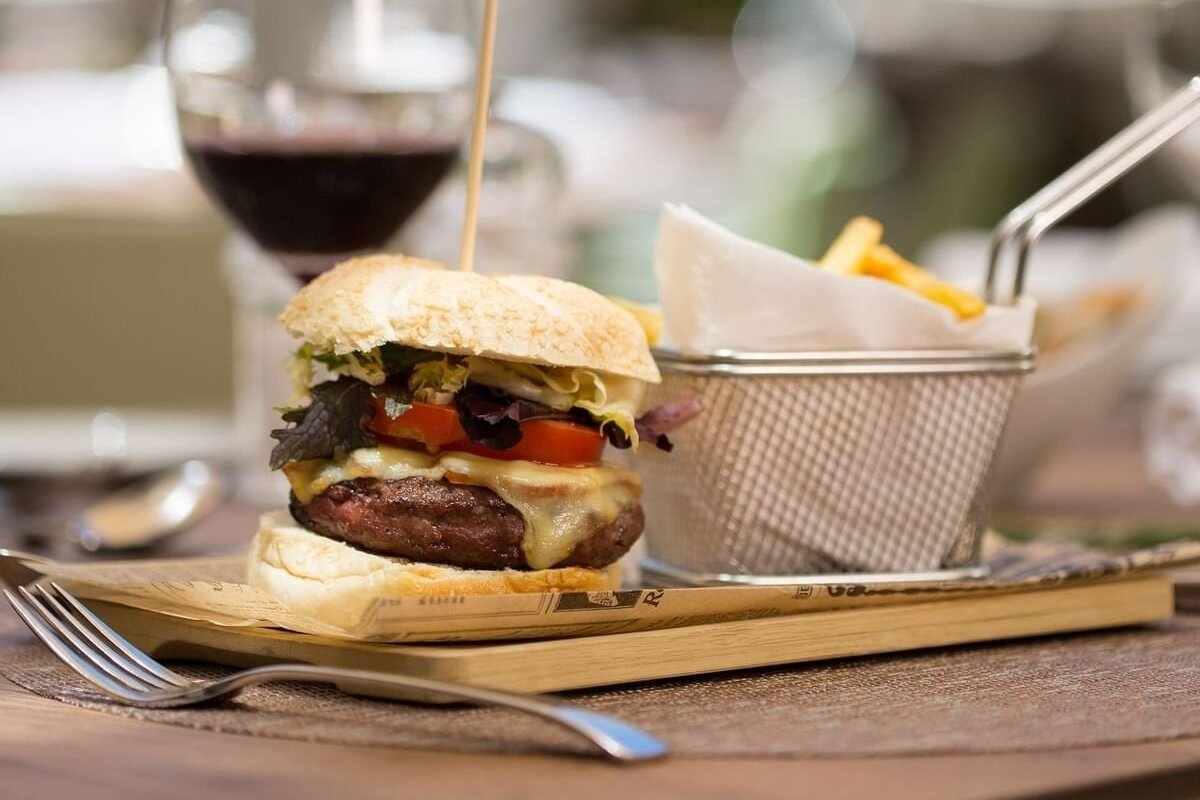 A gourmet cheeseburger, fries in a metal basket, a glass of red wine, and a fork are displayed on a wooden board, ready to be eaten.
