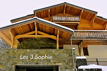 A multi-story wooden chalet, named 'Les 3 Sophie', features a stone base, balconies, and snow-dusted roofs, set against a winter backdrop.