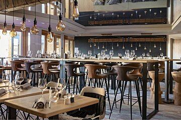 A modern, stylish restaurant interior features high bar tables with leather stools, set dining tables, and warm Edison bulb lighting creating an inviting atmosphere.