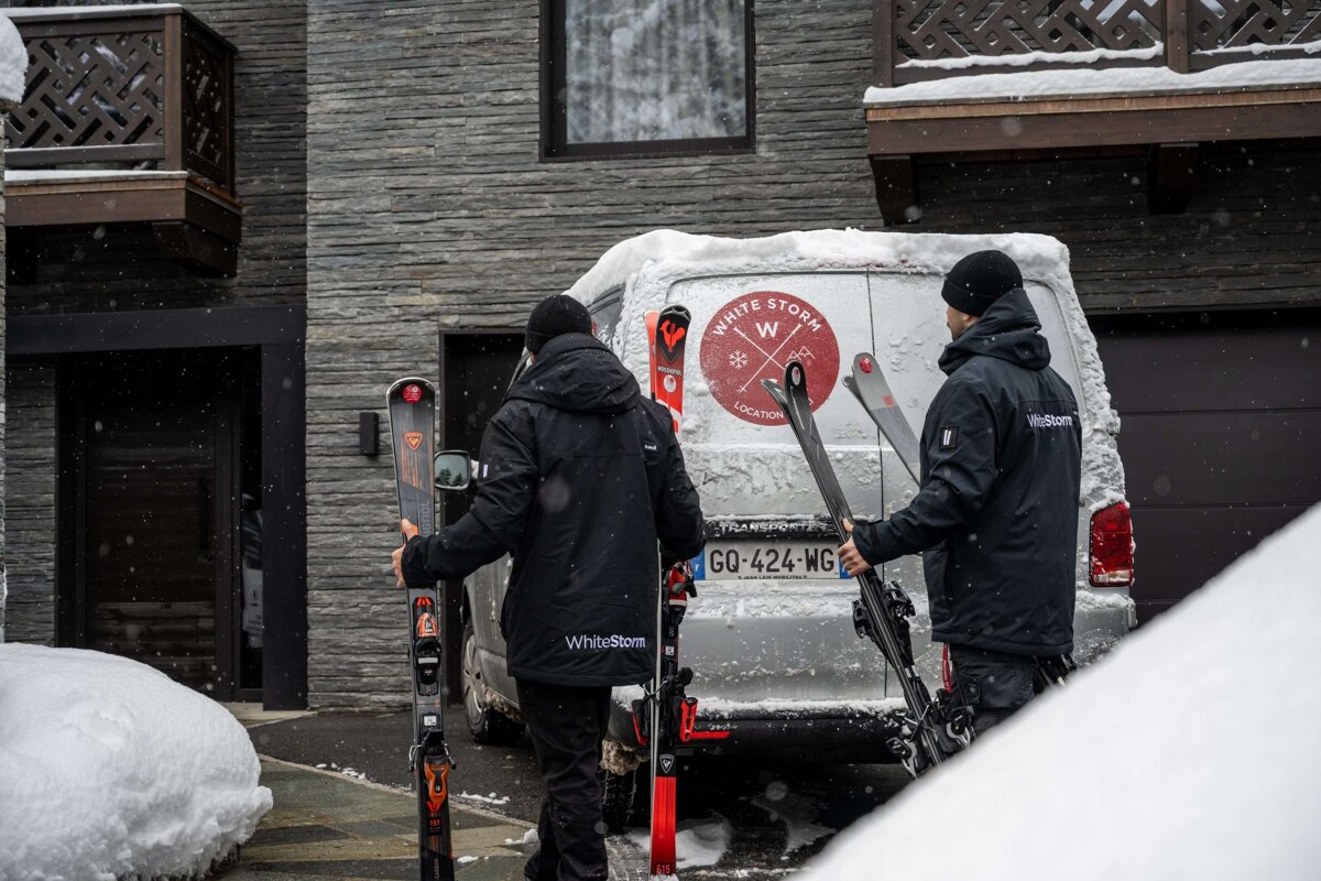 Two men carrying skis in front of a white storm van