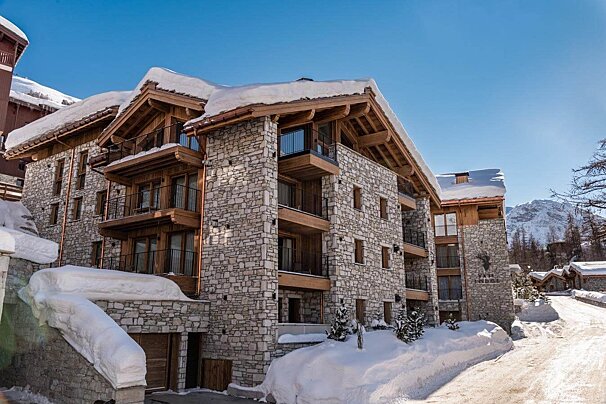 A large, snow-covered stone and wood alpine lodge with balconies, nestled in a winter mountain landscape under a clear blue sky.