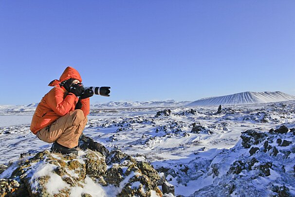 An orange-jacketed photographer crouches on snowy rocks, shooting a vast, wintry landscape with a distant snow-covered volcano under a clear blue sky.