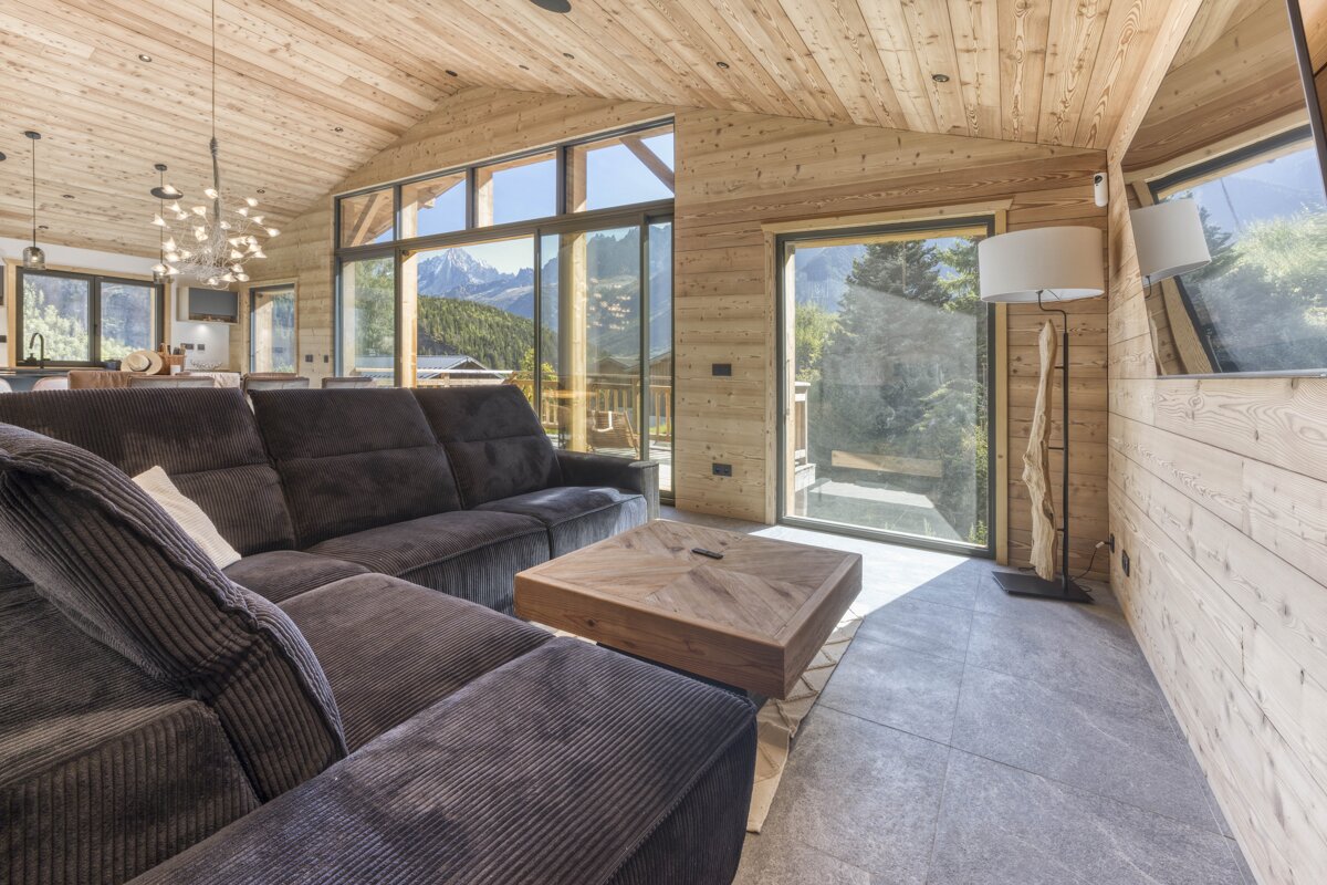 A modern, bright living room features extensive wood paneling, a large sectional sofa, and floor-to-ceiling windows with panoramic mountain views.