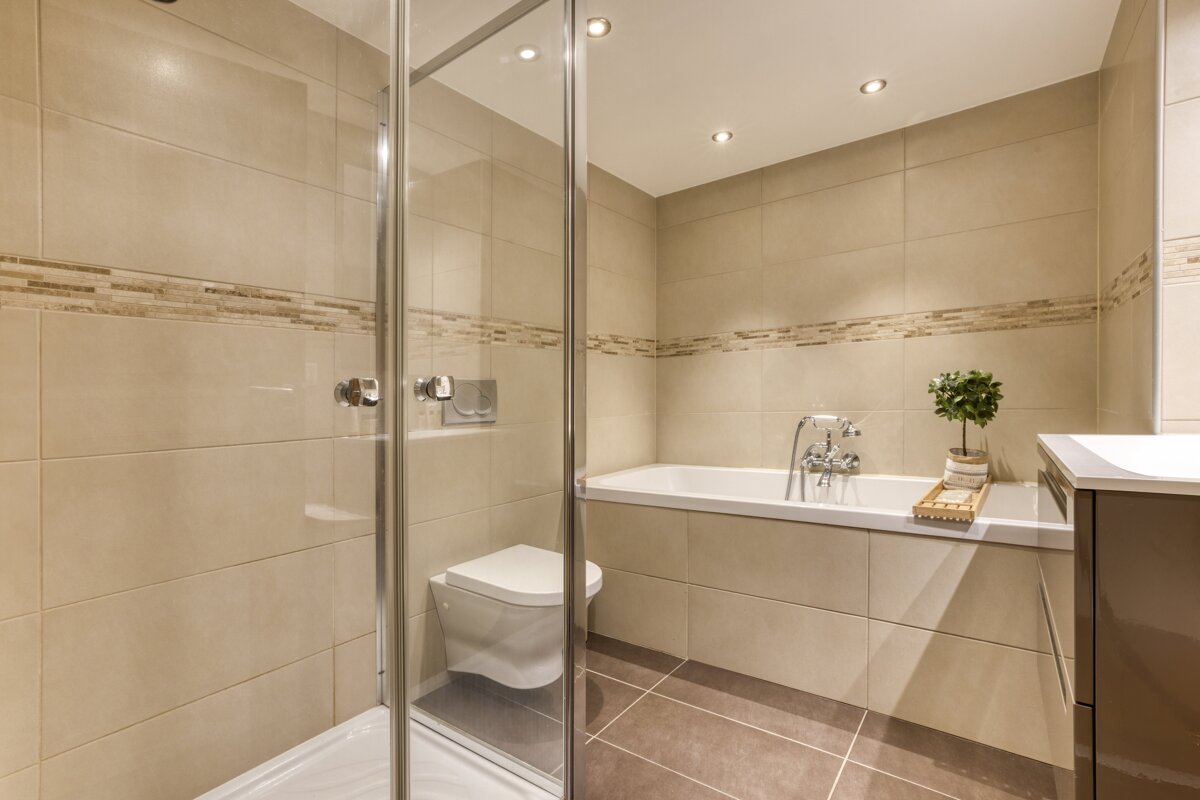 A modern, tiled bathroom features a glass shower enclosure with a toilet inside, a bathtub, and a vanity, all under recessed lighting.