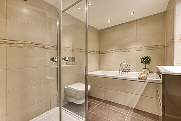 A modern, tiled bathroom features a glass shower enclosure with a toilet inside, a bathtub, and a vanity, all under recessed lighting.