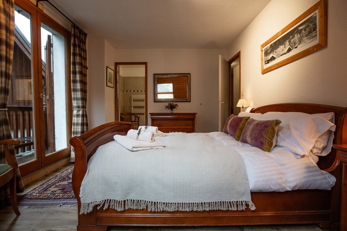 Cozy bedroom with a large wooden bed, patterned pillows, and a balcony. A bathroom is visible in the background through an open doorway.