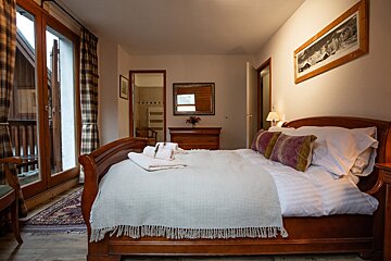 Cozy bedroom with a large wooden bed, patterned pillows, and a balcony. A bathroom is visible in the background through an open doorway.