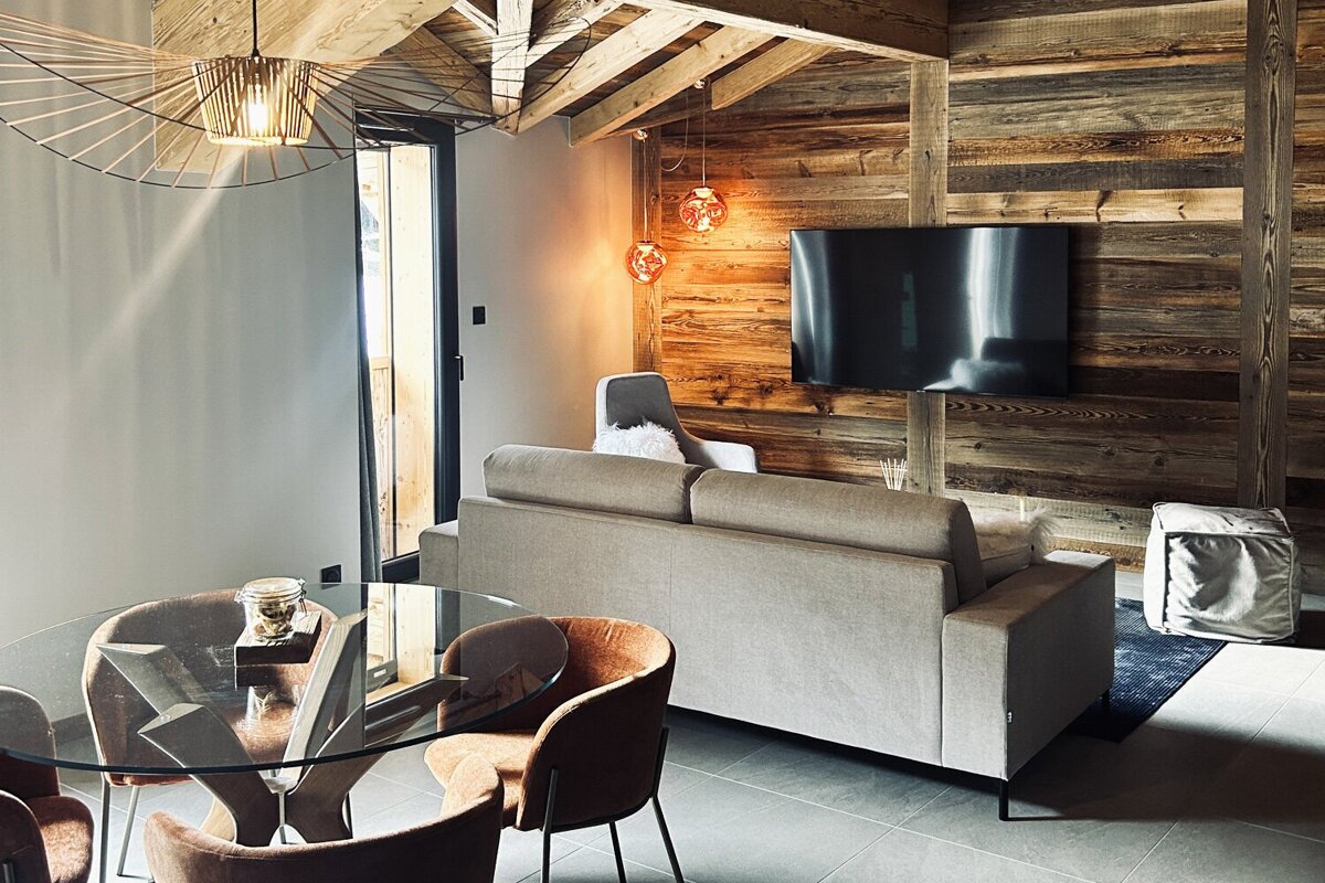 A modern chalet-style living and dining area featuring warm wood paneling, exposed beams, a comfortable sofa, glass table, and stylish lighting.