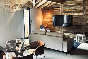 A modern chalet-style living and dining area featuring warm wood paneling, exposed beams, a comfortable sofa, glass table, and stylish lighting.