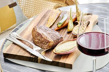 A steak and vegetables on a wooden cutting board next to a glass of wine