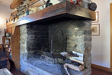 A rustic stone fireplace with a decorated wooden mantelpiece, featuring a cast-iron fireback, firewood, and a wooden built-in shelf.