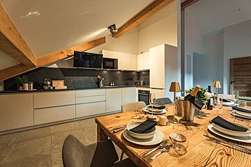 A contemporary kitchen with white cabinets, dark backsplash, and wooden beams. A wooden dining table set with black napkins, white dishes, and champagne awaits guests.