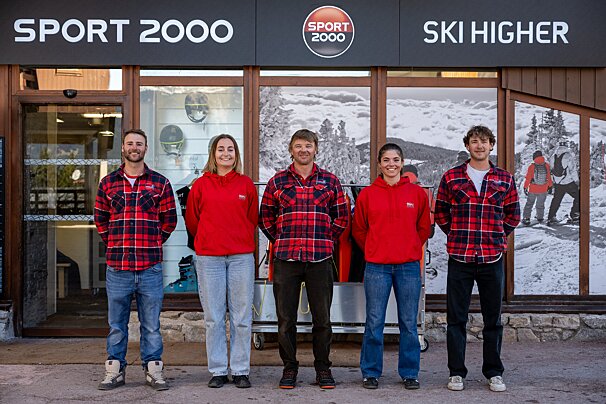 Five smiling staff members (3 men in plaid, 2 women in red hoodies) stand before a 'SPORT 2000' store, with winter scenes and gear in the windows.