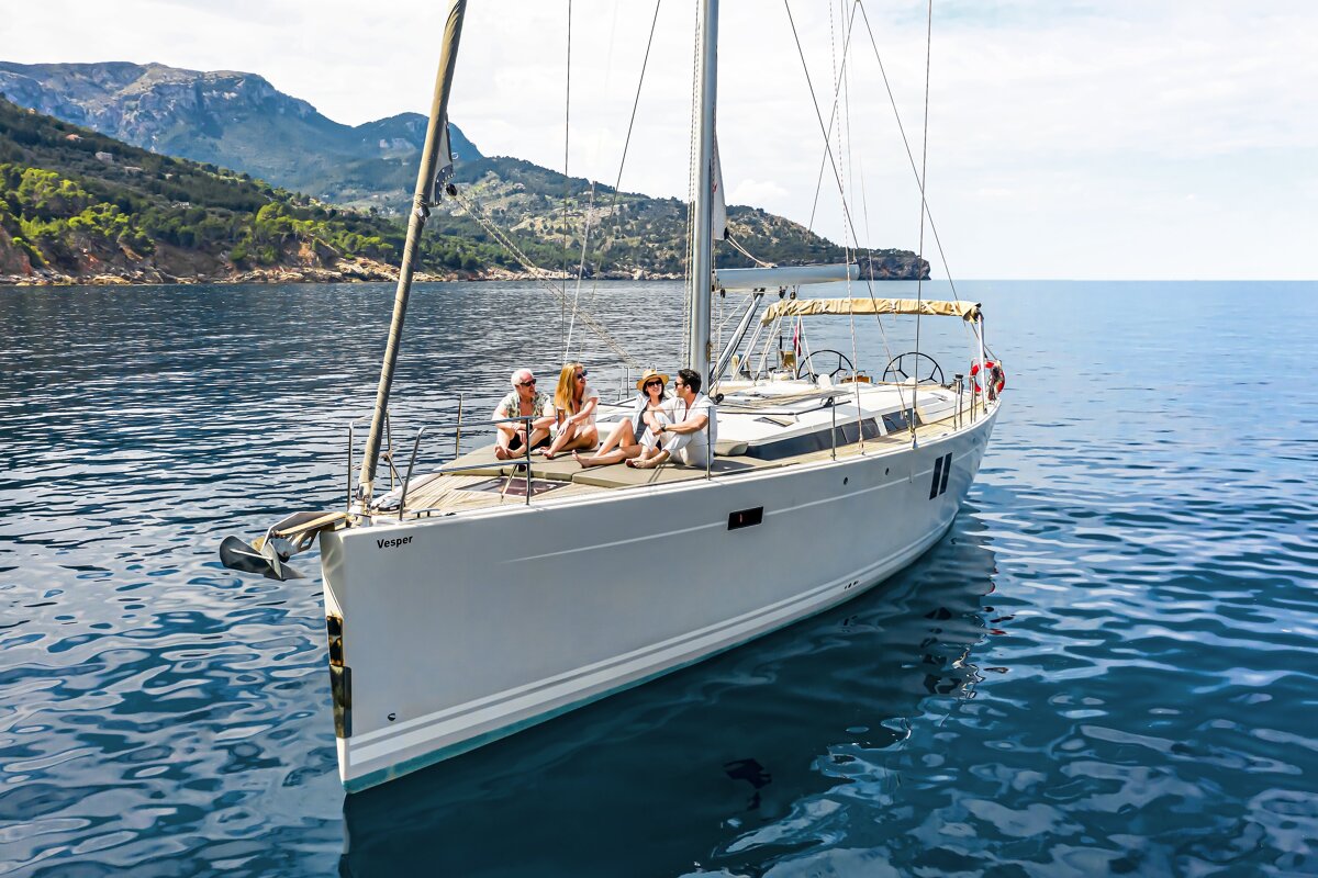 Four people relax on the deck of a sailboat named Vesper in calm, blue waters, with mountainous cliffs in the background.