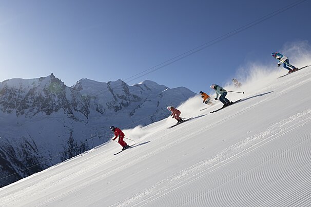 Five skiers descend a sunny, groomed snow slope, kicking up powder, with majestic snow-capped mountains and a clear blue sky beyond.