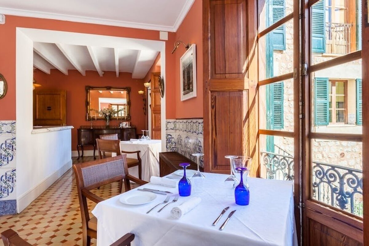 A cozy, rustic restaurant with terracotta walls, set tables with blue glasses, and a window offering a view of buildings with green shutters.
