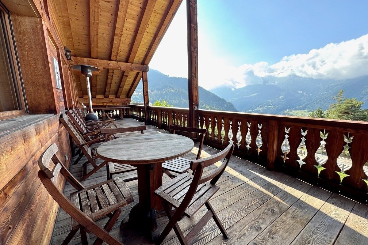 Sunny wooden balcony with tables and chairs, offering panoramic views of a lush mountain valley and cloudy sky.