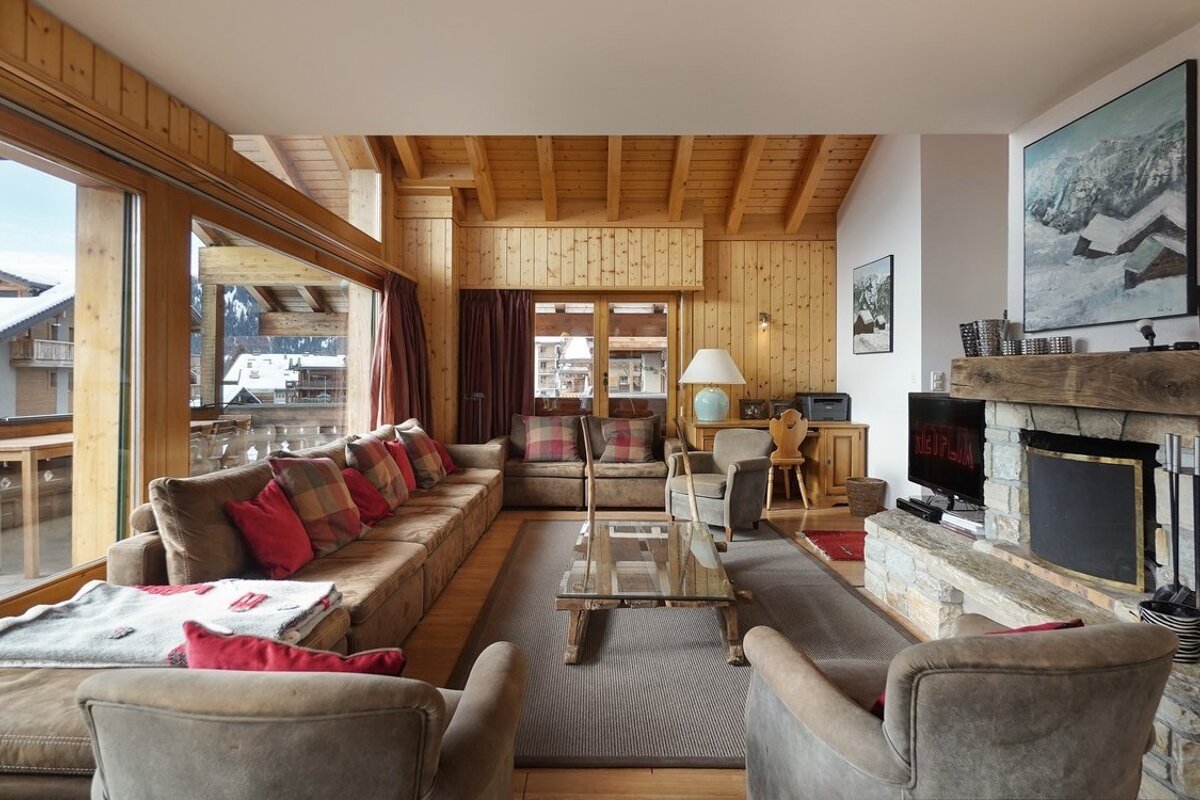 A warm, wood-paneled chalet living room with a large sofa, stone fireplace, and wide windows revealing snowy peaks and buildings.