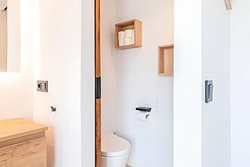 A clean, modern toilet area with white walls, wooden details, open shelves holding toilet paper, and stylish fixtures.