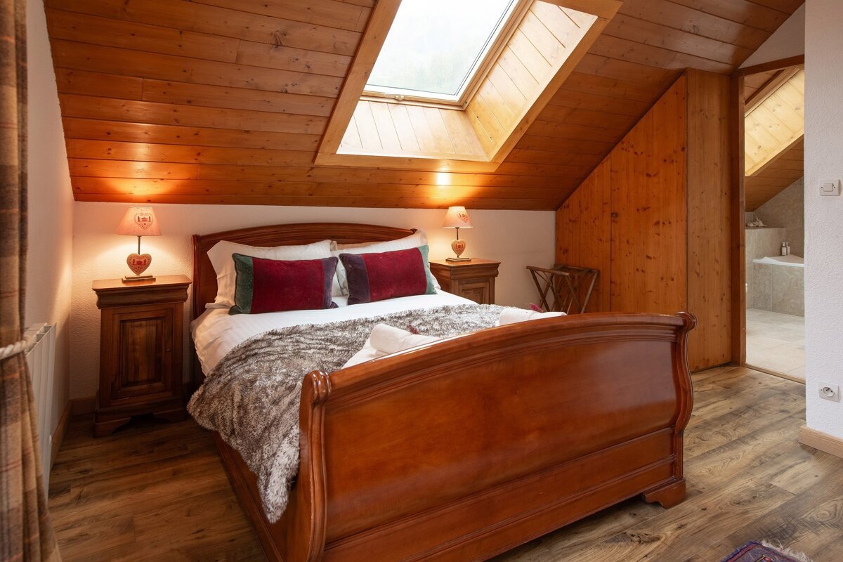 A cozy attic bedroom featuring a wooden sleigh bed, red pillows, and a faux fur throw. A slanted wooden ceiling with a skylight and rustic nightstands create a warm ambiance.